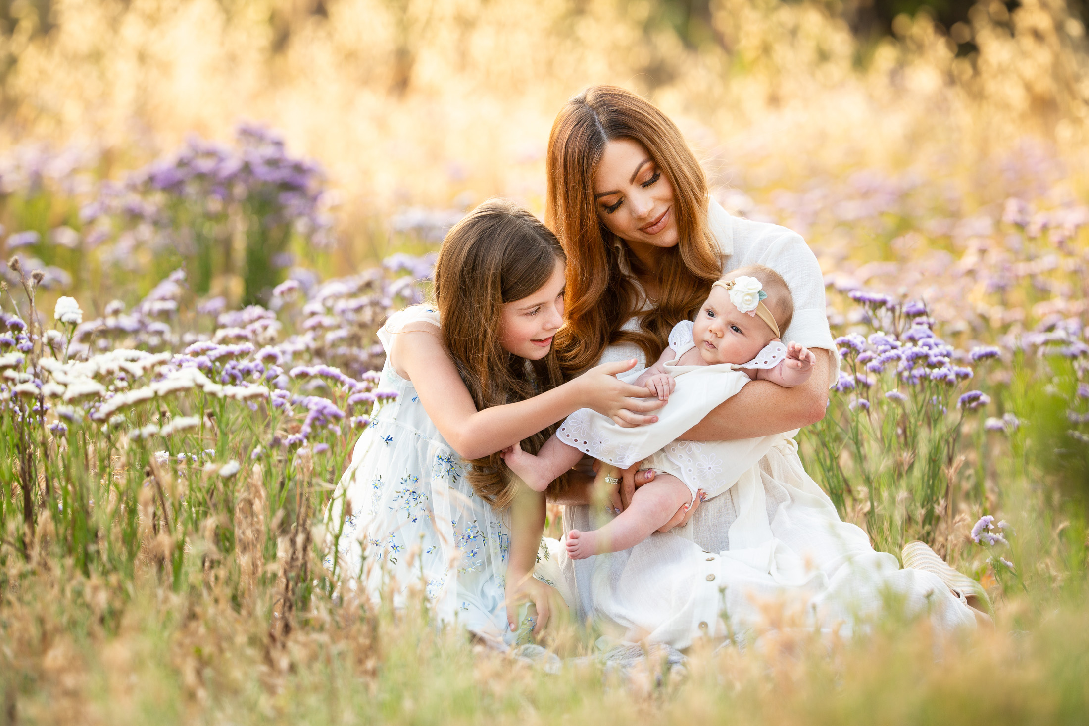 photo of a mom, daughter and young baby girl. they are all dressed in white and sitting in a purple-flower field
