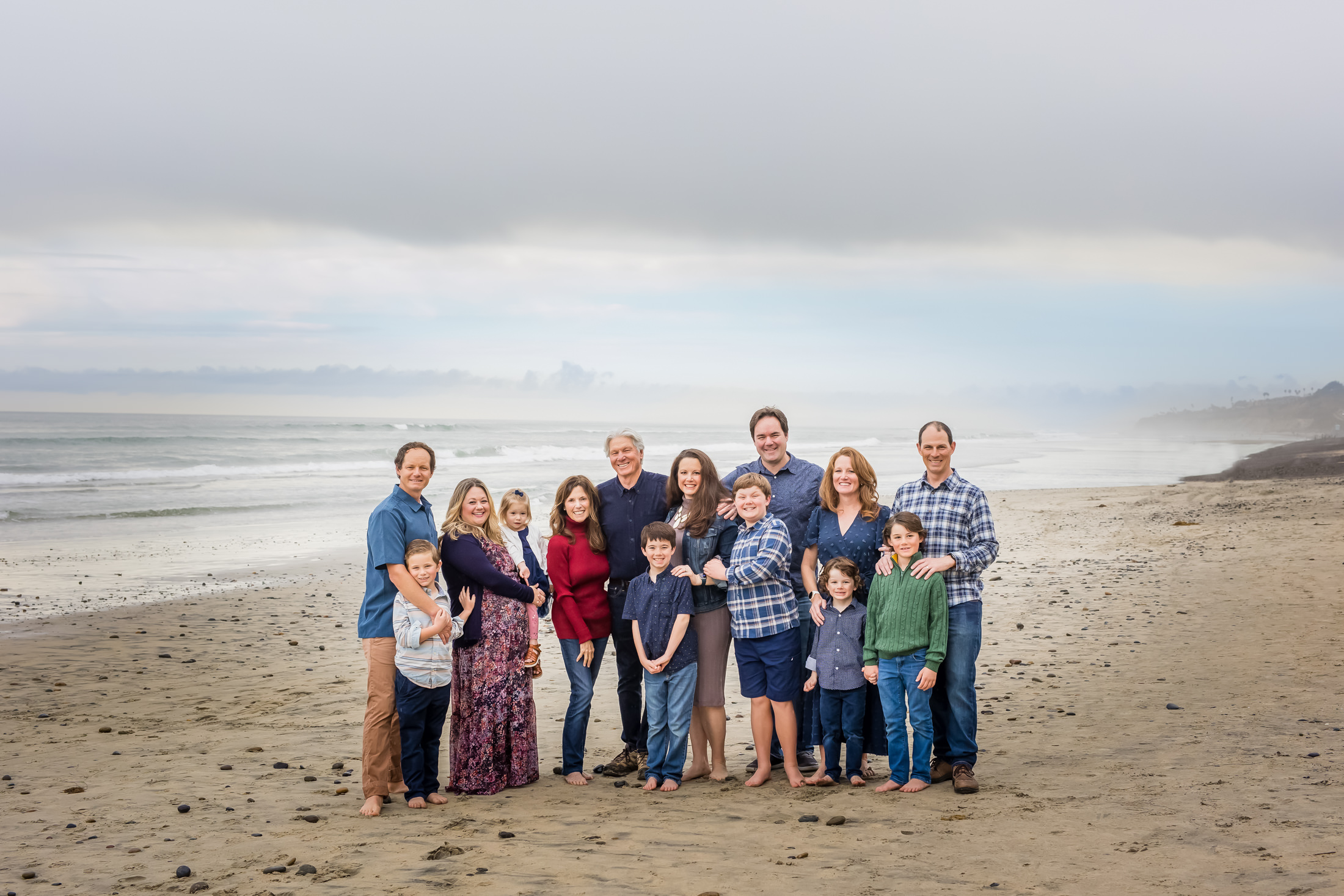 portrait of a family of 14 barefoot on the sand at low tide at the beach at sunset. colors are blue, green and red.