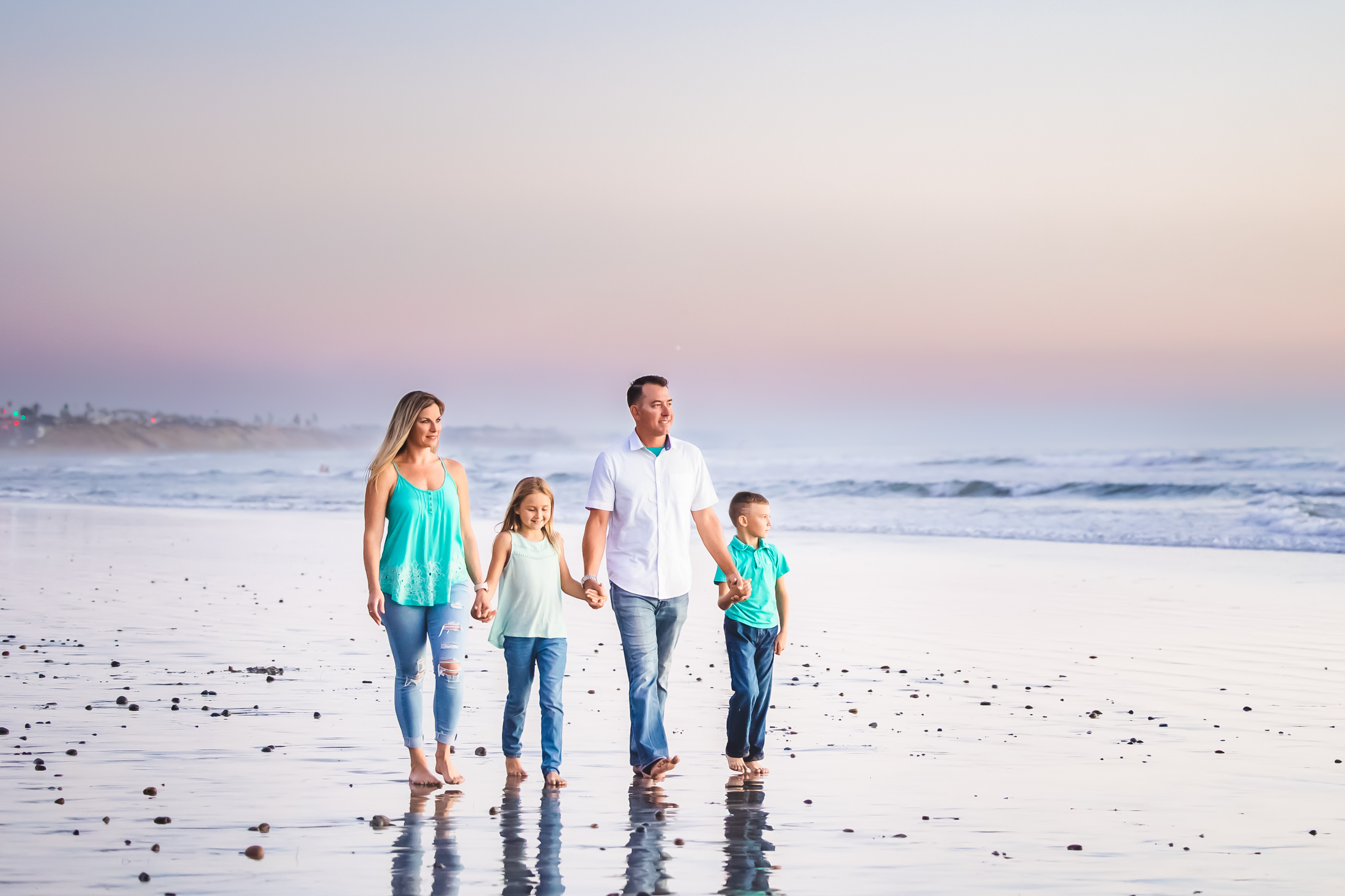 portrait of a family of 4 holding hands and walking along the edge of low tide at the beach at sunset