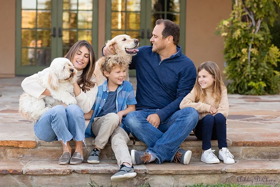 photo of a family of four. mom, dad, and two kids. they are seated and cuddling with their two dogs