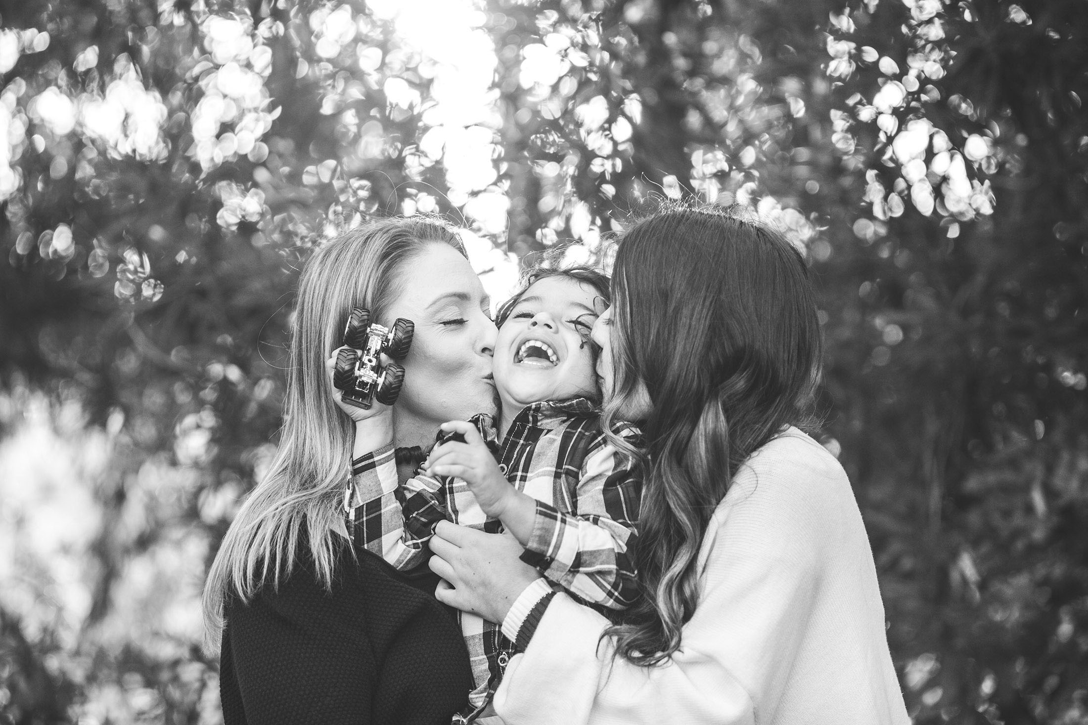 black and white photo of two women hugging and kissing a young boy while he laughs and holds his toy truck