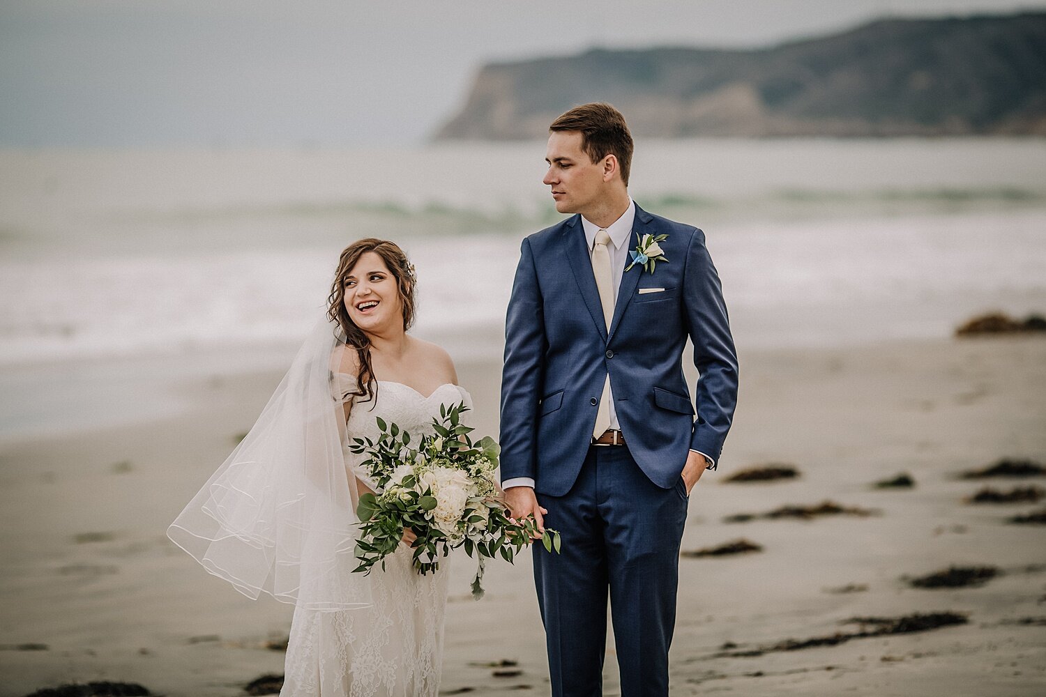 photo of a bride and groom holding hands and standing on the stand at the beach with point loma and the ocean in the background